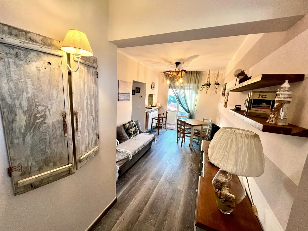 Il soggiorno di Shabby House a Forte dei Marmi: divano con cuscini, corridoio con mensole in legno sbiancato e tavolo da pranzo davanti alla porta-finestra